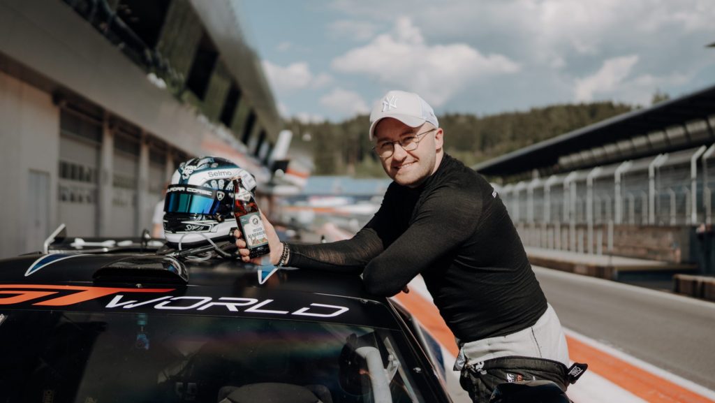 Ein Fahrer lehnt mit einer Flasche Schnaps in der Hand an einem Rennwagen, daneben steht ein Rennhelm und im Hintergrund ist eine Rennstrecke zu sehen.